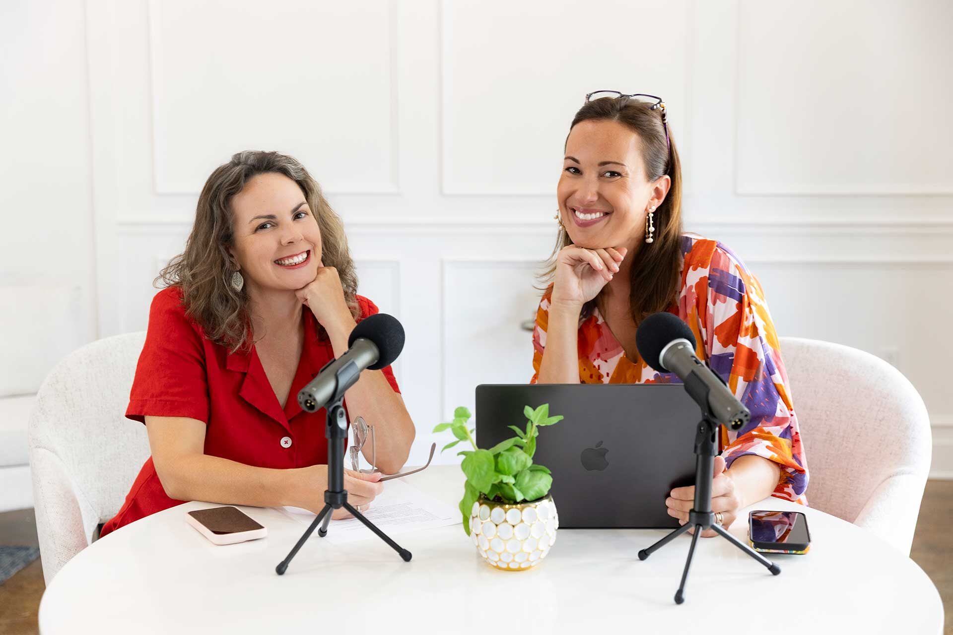 Two smiling women sit at a round white table with microphones, a laptop, a phone, and a potted plant.