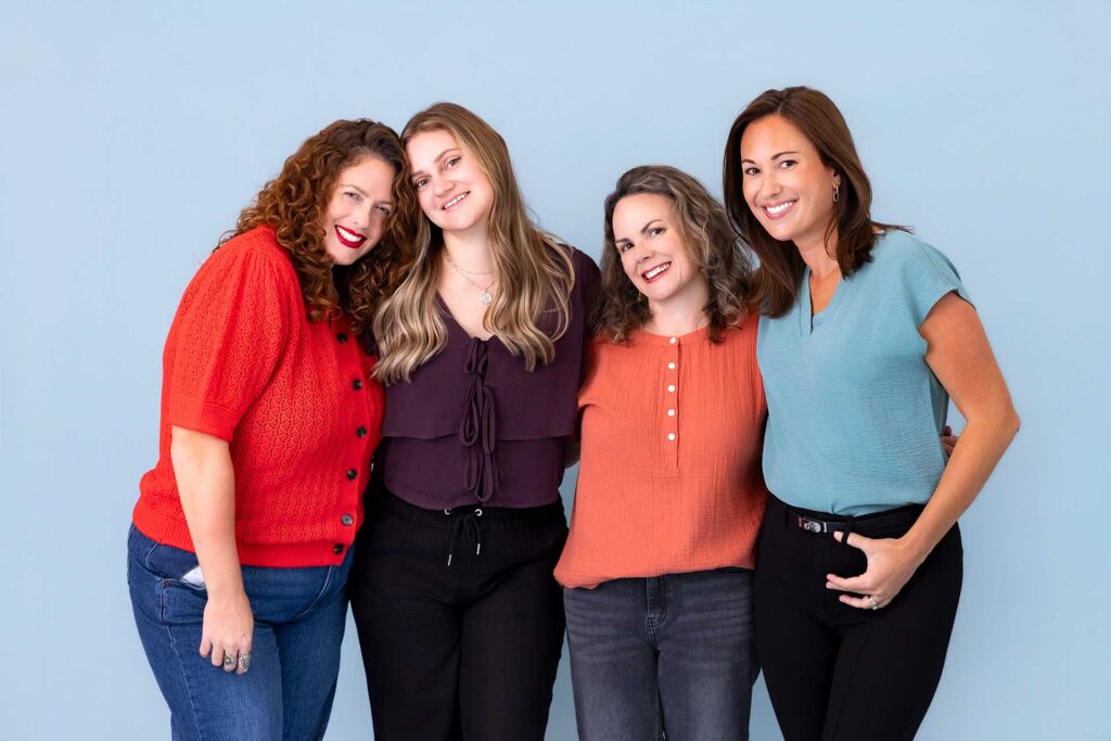 Four women stand together smiling in casual clothes against a light blue background.