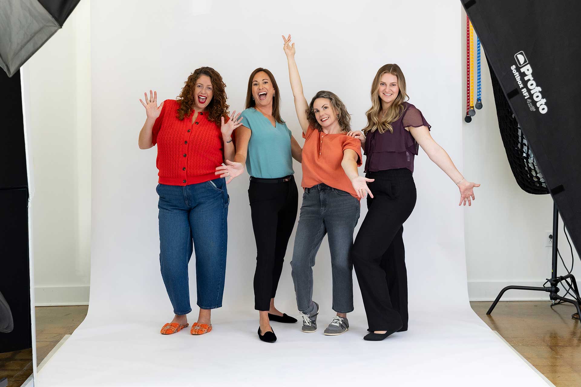 Four smiling women pose playfully in a photo studio with white background and professional lighting.
