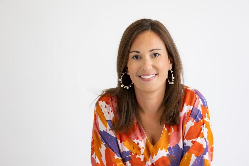 Woman with straight brown hair wears large hoop earrings and a colorful patterned blouse, smiling at the camera against a plain white background.