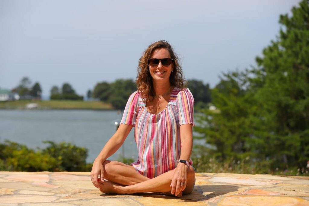Woman with wavy hair and sunglasses sits cross-legged on a stone patio, smiling. She wears a striped shirt and shorts.