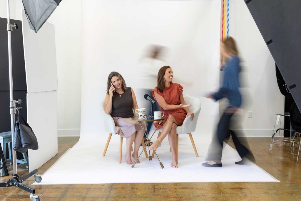 Two women sit chatting on chairs with coffee, while two blurred people walk by on a white photo set.
