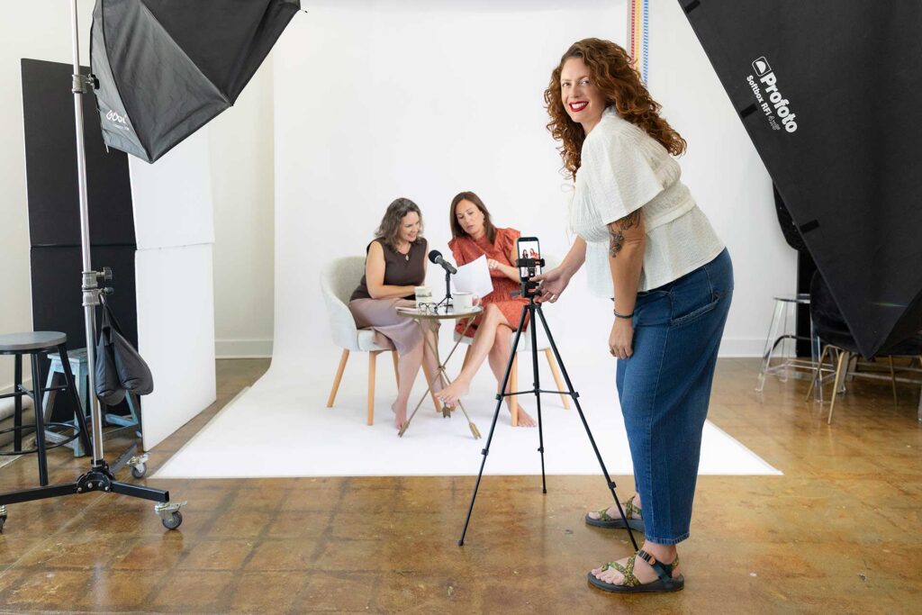 Three women in a photo studio; two record at a table, one smiles at the camera, holding a phone on a tripod.
