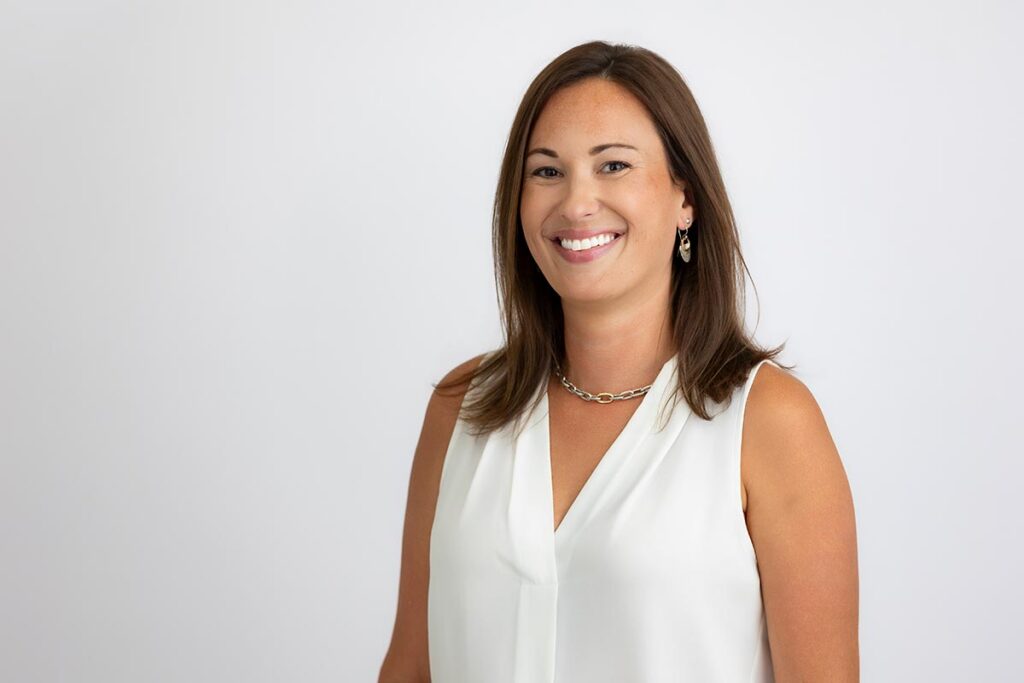 Woman with straight brown hair, wearing a sleeveless white blouse and gold necklace, standing and smiling against a plain white background.