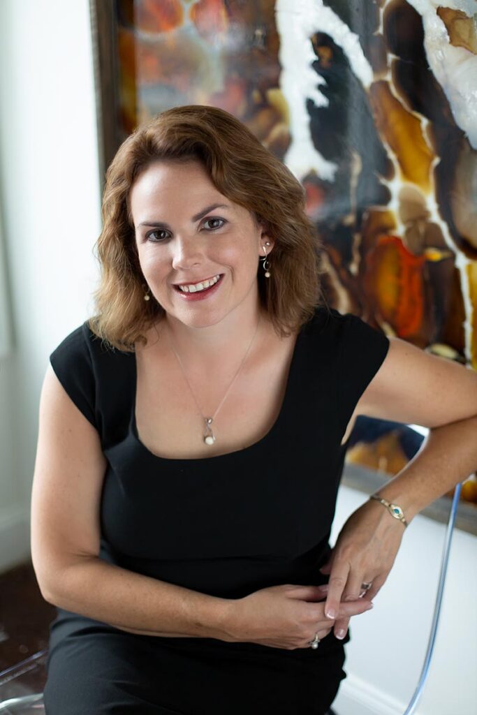 Woman with shoulder-length brown hair wearing a black dress, smiling and sitting in front of an abstract painting.