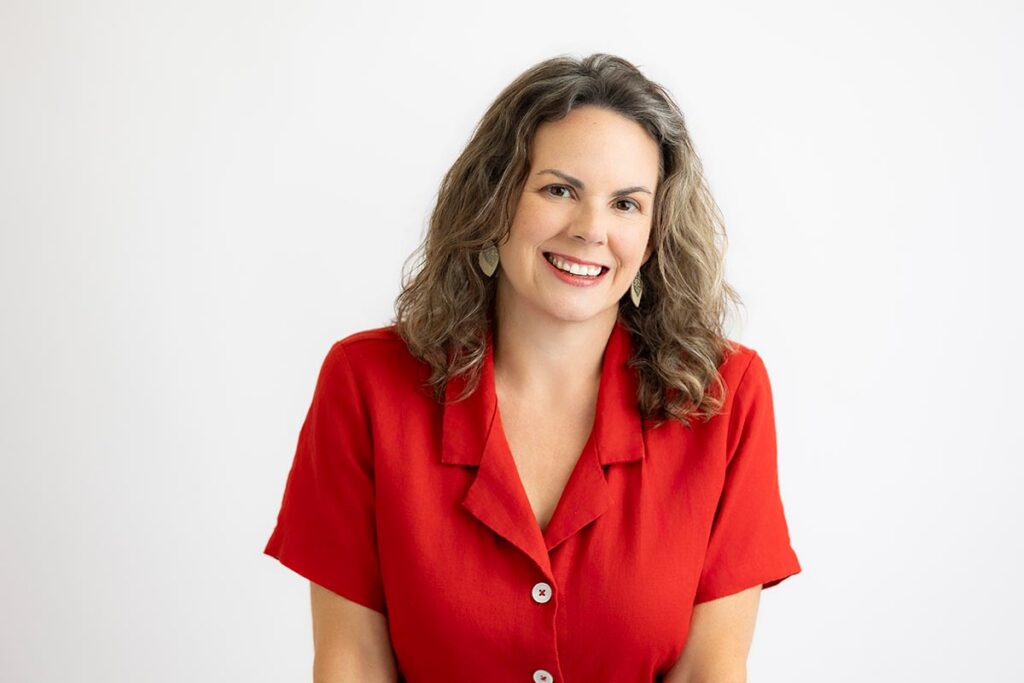 Woman with wavy brown hair wearing a red button-up shirt smiles at the camera against a plain white background.