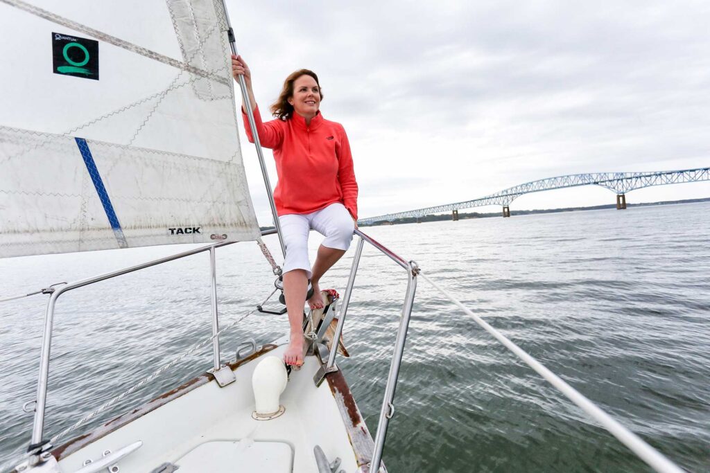 Woman in a red top sitting on the bow of a sailboat with a bridge and water in the background.