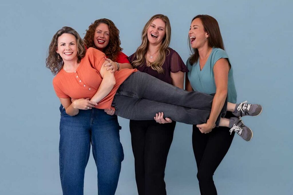 Four women smile and laugh while two of them lift another woman horizontally in front of a blue backdrop.