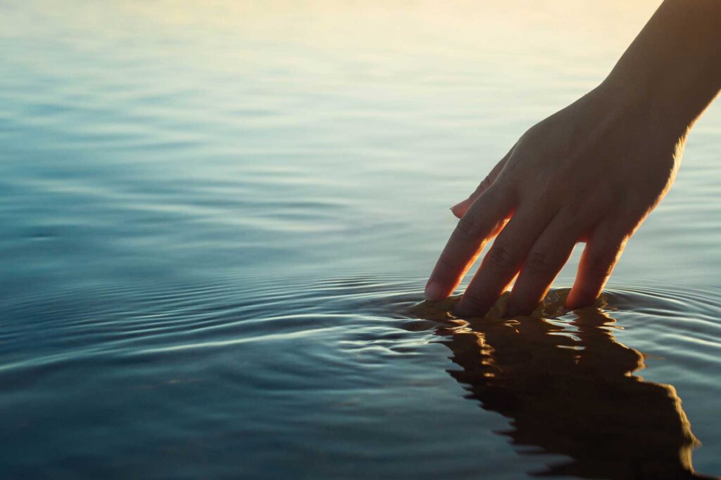 A hand touching the ocean water causing ripples in front of a beautiful sunset.