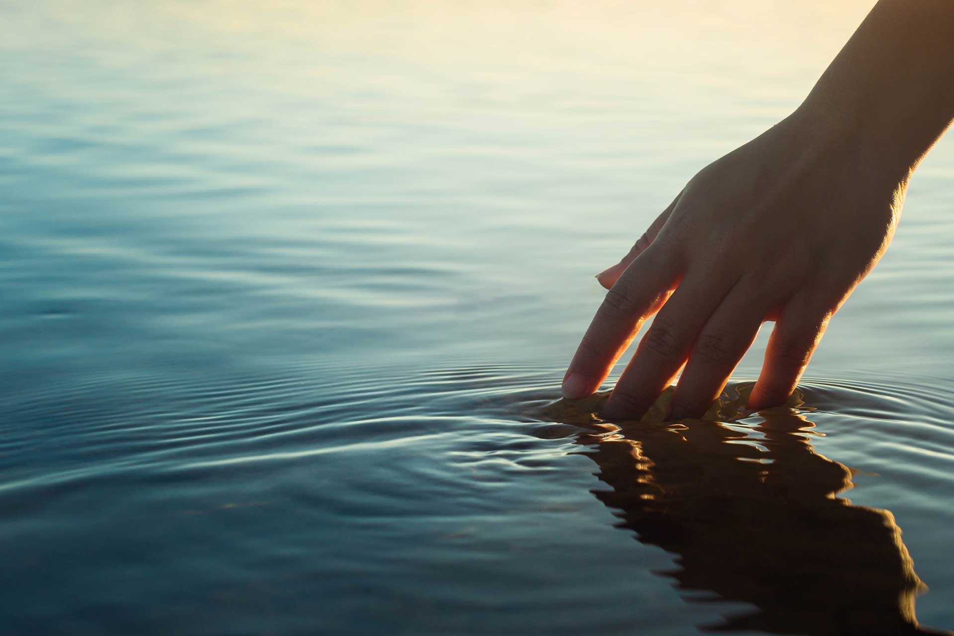 A hand touching the ocean water causing ripples in front of a beautiful sunset.
