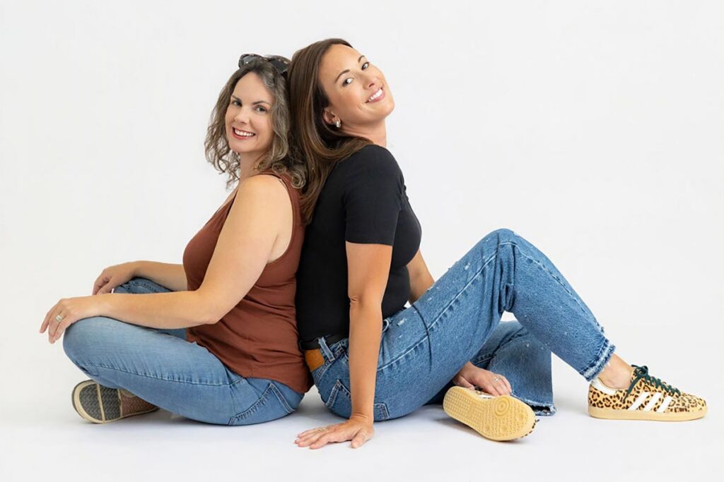 Two women sit back to back on the floor, smiling, in casual tops and jeans against a white background.