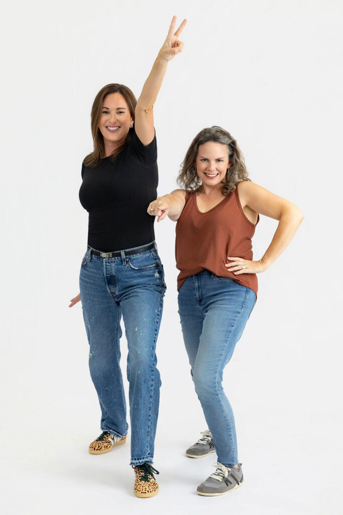 Two women in casual clothes pose playfully, one making a peace sign and the other pointing forward.
