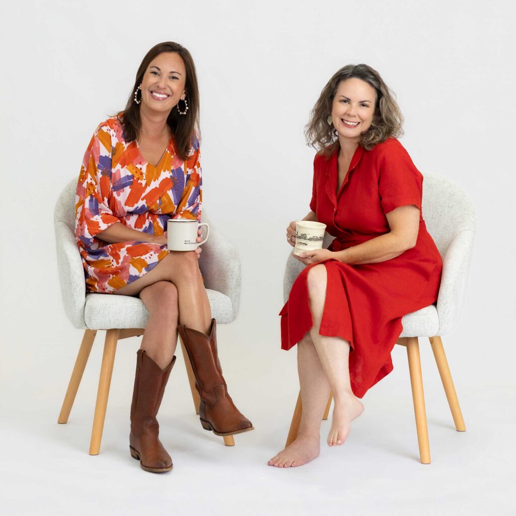 Two women smiling, sitting on chairs with mugs; one wears boots, the other is barefoot in a red dress.