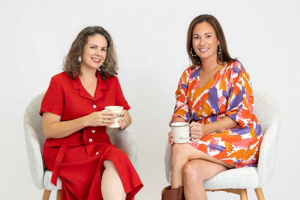 Two women sit on chairs, clink mugs, and smile with a microphone and glasses on the small table between them.
