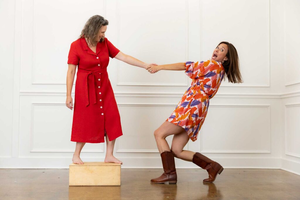 Two women, one barefoot on a box in red dress, holding hands with another in boots and a patterned dress.