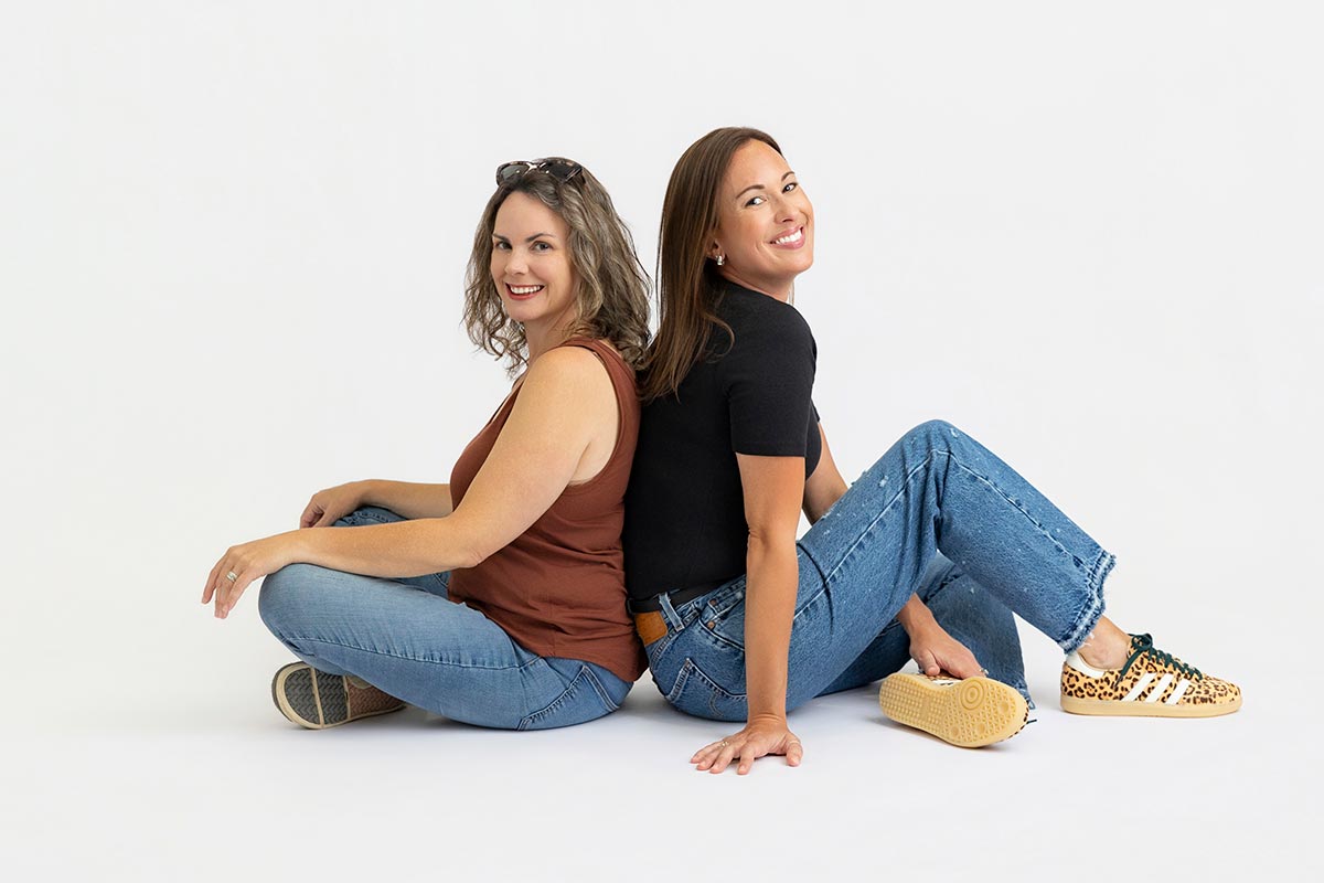 Two women sit back-to-back on the floor, smiling, in casual jeans and tops against a plain white background.