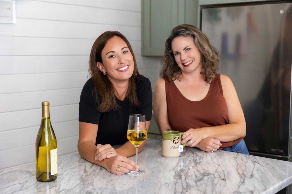 Two women smiling at a kitchen counter with a wine glass, bottle, and coffee mug.