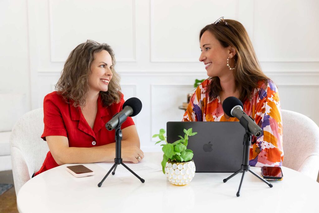 Two women sit at a table with microphones, a laptop, and a plant, smiling at each other.