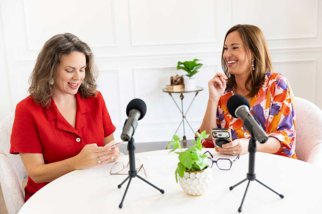 Two women sit at a round table with microphones, smiling and looking at their phones.