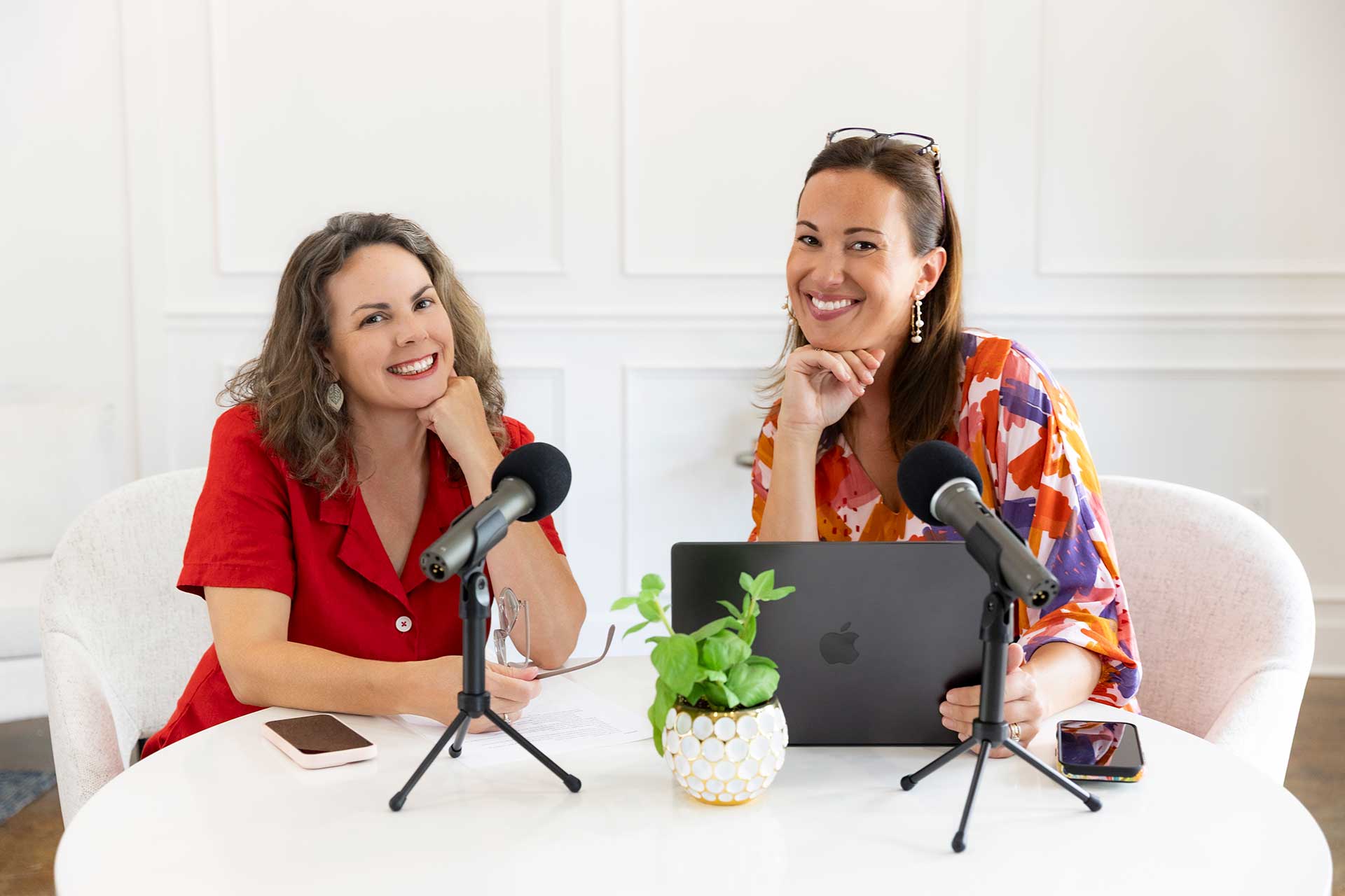 Two smiling women sit at a round white table with microphones, a laptop, a phone, and a potted plant.