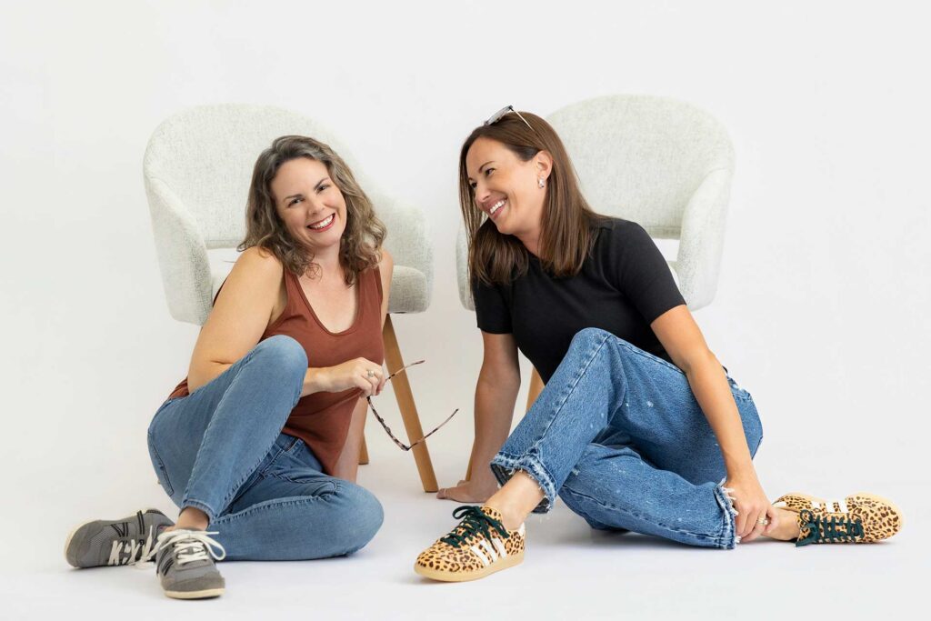 Two women sit on the floor smiling and laughing, with white chairs behind them against a white background.