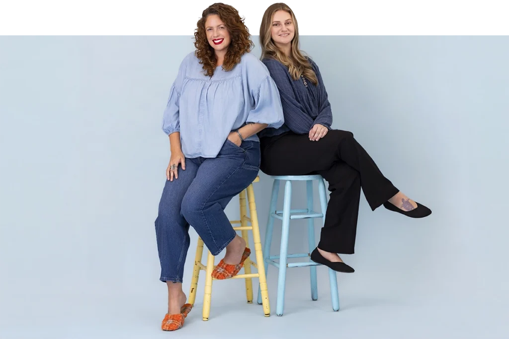 Two women standing side by side, smiling, wearing loose blue tops and casual pants against a plain background.