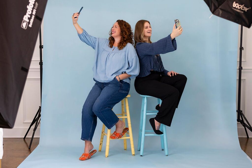 Two women sit back to back on stools, smiling and taking selfies in a photography studio with blue background.