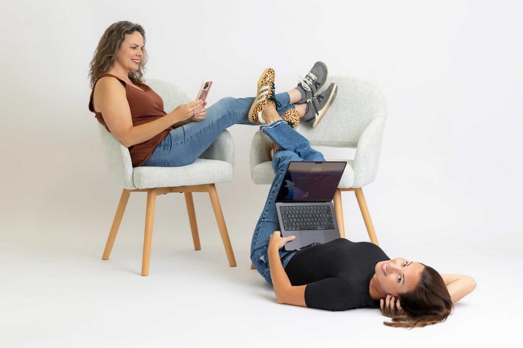 Two women work together, one sitting in a chair on her phone, the other lying on the floor using a laptop.