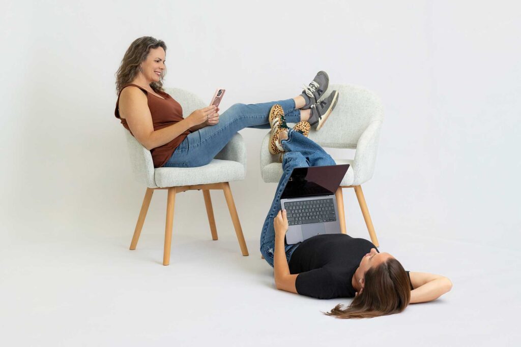 Two women relaxing, one in chair on phone, other on floor with laptop, legs resting on chair’s arm.